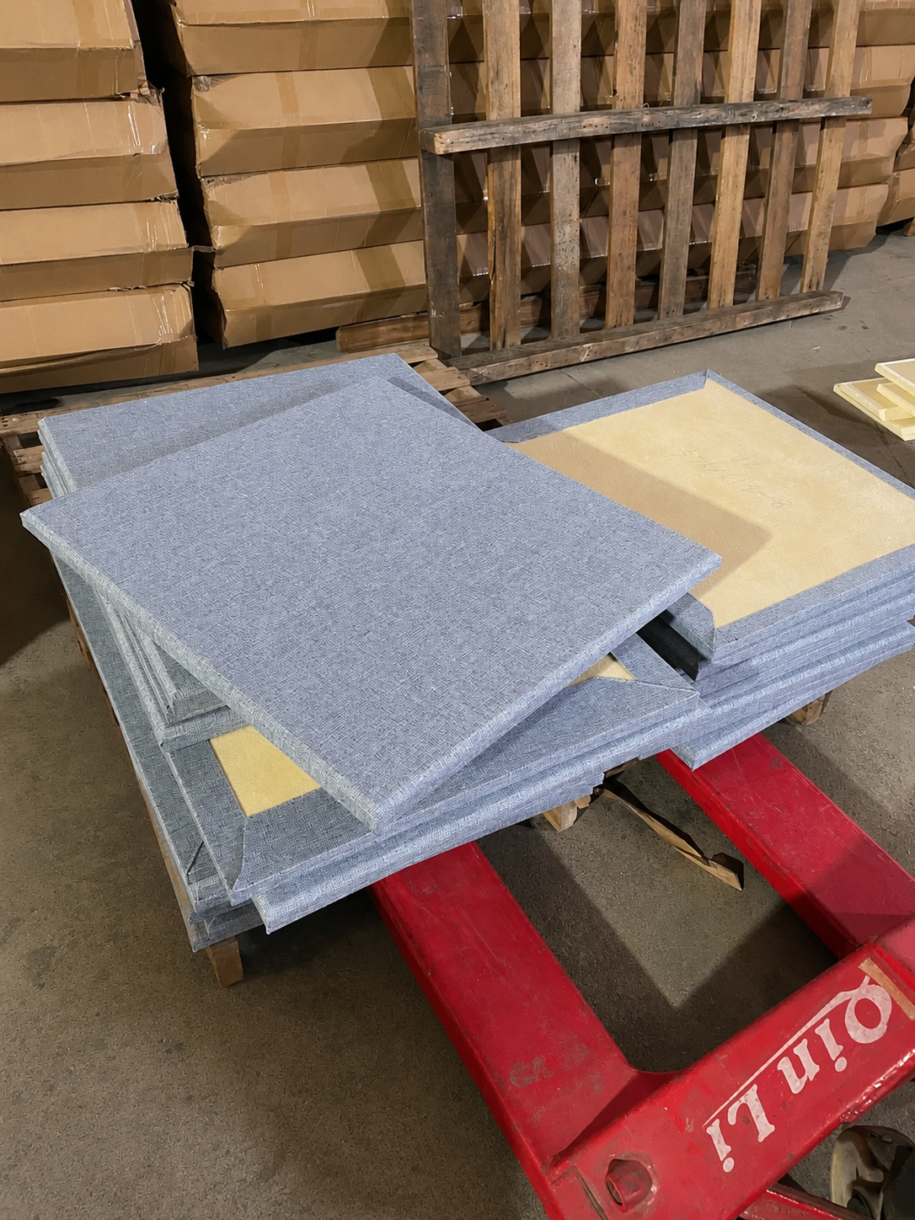 A stack of fabric-wrapped fiberglass acoustic panels on a pallet jack in a manufacturing facility, showing the grey fabric finish and internal yellow sound-absorbing core.