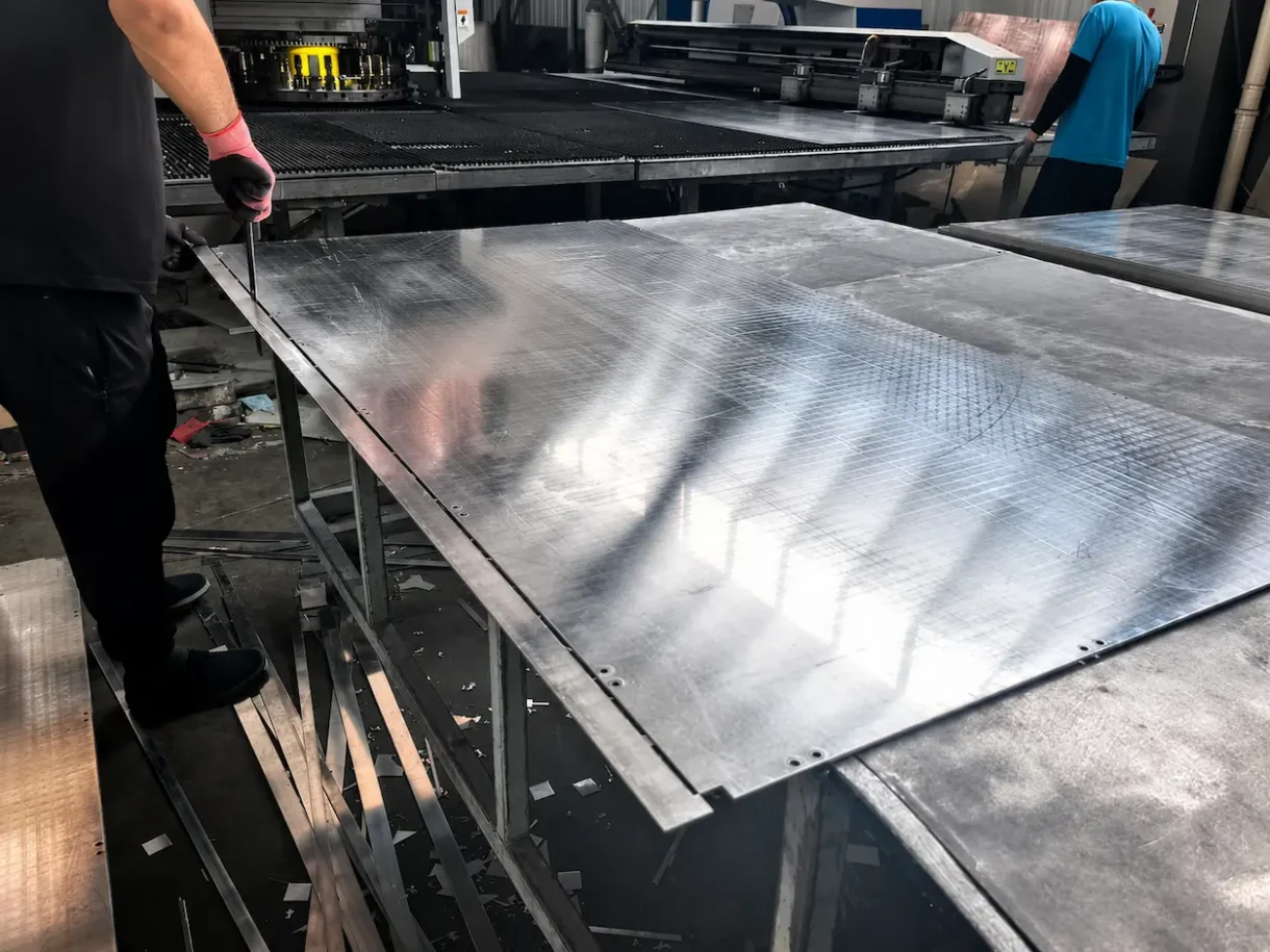 Workers processing a large aluminum sheet on factory fabrication equipment